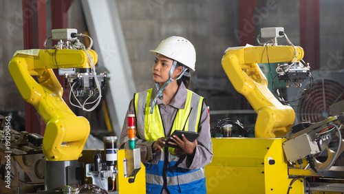 Industrial plants factory technician robot automation safety helmet tablet inspection in industrial floor as focused worker guides yellow robotic arm inside modern production area
