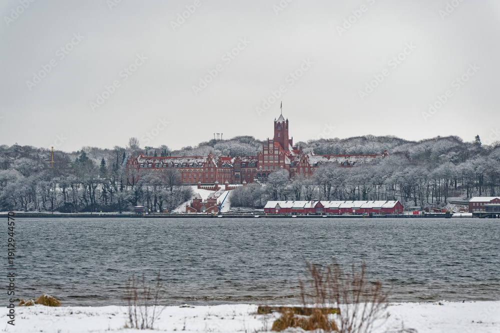Fototapeta premium Historic red brick building surrounded by snowy trees near a calm body of water in Flensburg