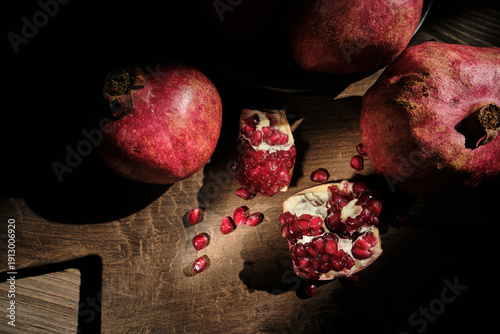 Flat lay photography of fresh pomegranates in a bowl and broken pieces with red seeds on a rustic cutting board, featuring artistic natural shadows on a wooden background.