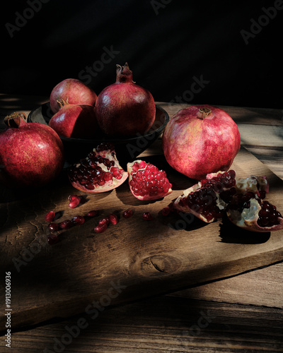 A dark, atmospheric still life featuring whole and opened pomegranates with juicy red seeds scattered on a wooden table, highlighted by dramatic directional light against a black background.