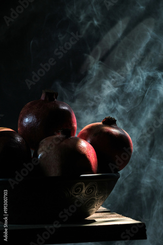 A vertical professional shot of pomegranates in a bowl and sliced pieces on a rustic wood surface, featuring ethereal light beams and smoke swirls against a dark moody background.