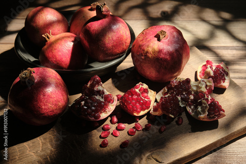 Flat lay photography of fresh pomegranates in a bowl and broken pieces with red seeds on a rustic cutting board, featuring artistic natural shadows on a wooden background.