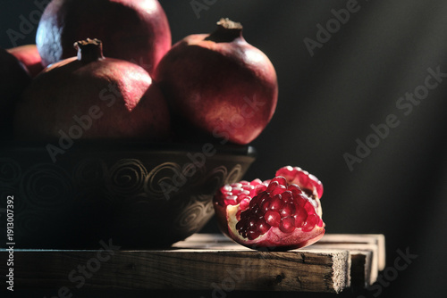 A close-up shot of a broken pomegranate piece showing glistening red seeds in sharp focus, with whole pomegranates in a bowl and volumetric light rays in the blurry background