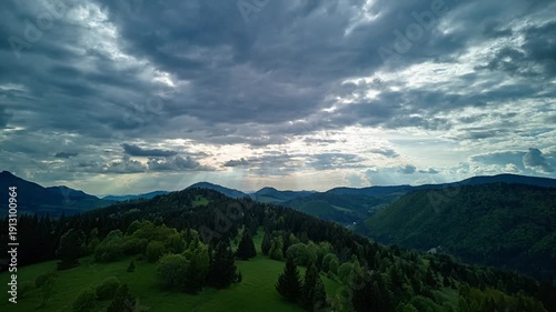 Aerial drone hyperlapse of moving clouds and sunbeams over mountain forest and meadows.