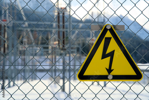 Triangular yellow high-voltage danger sign on fence, warning for electric shock, power transformer station out of focus behind, electrical safety symbol, utility infrastructure