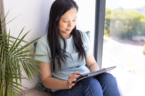 Woman sitting on cushioned window seat wearing blue top and dark jeans, holding tablet, plant left