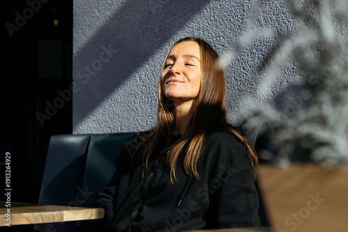 Tableau sur toile Young woman with long hair, wearing a black hoodie, enjoys sunlight while sittin