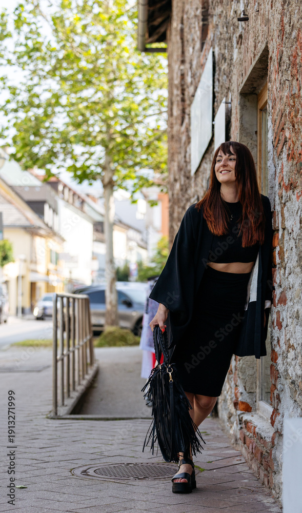 Αυτοκόλλητο Stylish woman in black outfit with fringe bag stands against a rustic stone wall