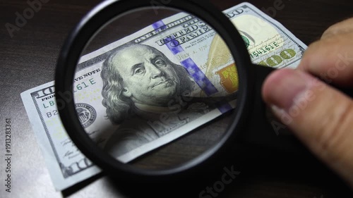 Examination of american dollar (USD) cash money with magnfication glass, loupe. Concept of authenticity check, fight against counterfeiting of banknotes, anti fraud measures in USA. Close up view.