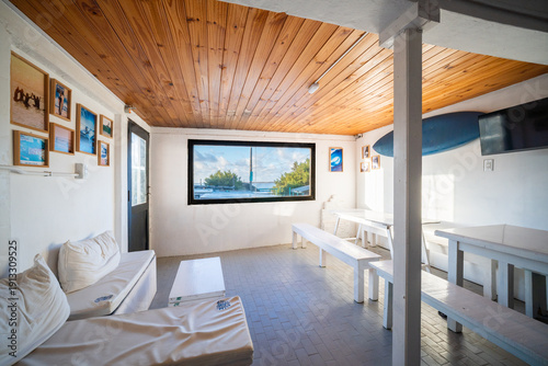 A bright, coastal-style lounge features white furniture, wooden ceilings, and surf-themed decor. Large windows offer a stunning view of the beach and ocean in a relaxing atmosphere.