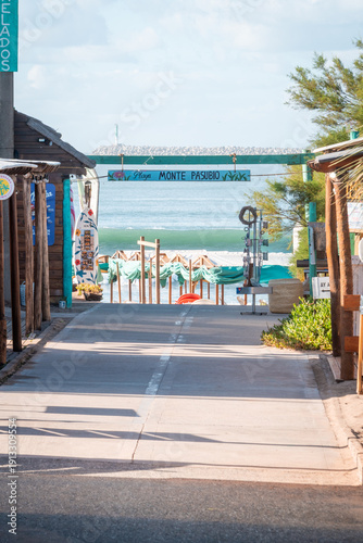A scenic entrance to Monte Pasubio beach features a paved path leading to the turquoise ocean, framed by rustic wooden structures and a decorative surfboard under a clear sky.