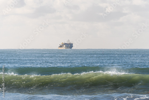 A large cargo ship sits on the calm horizon of a blue ocean, with a gentle green wave breaking in the foreground under a soft, hazy sky. A peaceful maritime scene