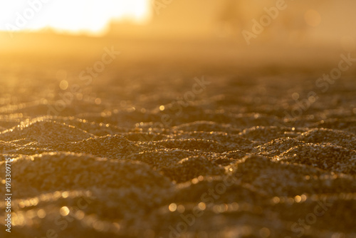 Golden sunset light reflecting over wet sand, capturing delicate textures and shimmering highlights in a calm, minimal coastal scene.