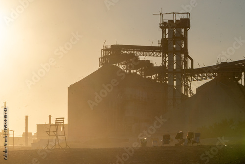 Industrial plant silhouetted at sunrise, with steel structures and dust in the air glowing under warm golden light, capturing a raw and powerful industrial scene.