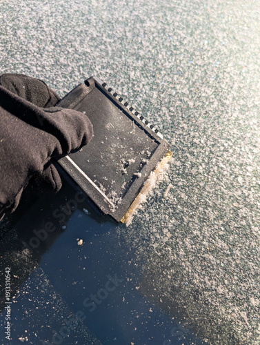 Close up of a gloved hand using a black plastic scraper to remove thick white frost from a car windshield