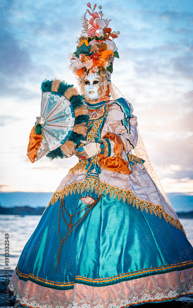 Fototapeta premium Venice, Italy - February 09, 2026: Elegant masked figure in ornate costume holds a fan at the Venice Carnival, showcasing intricate details and vibrant colors against a scenic backdrop