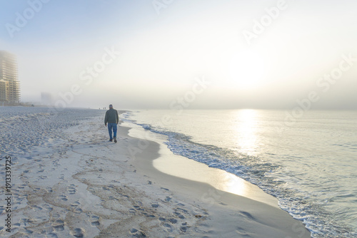 A man in warm clothes walking on a foggy beach at sunrise.