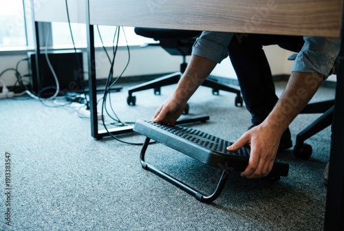 Office worker adjusting a black ergonomic footrest under a desk for better posture and comfort while sitting.