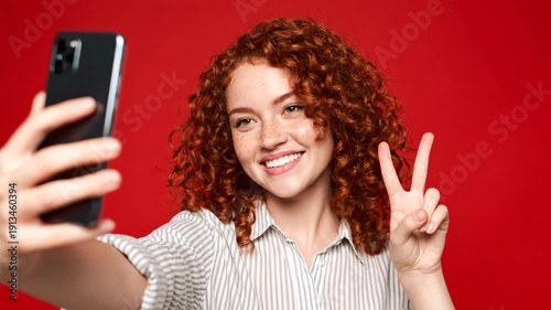 A young woman with curly red hair and freckles takes a selfie with her phone, making a peace sign and puckering her lips against a vibrant red background. She wears a striped shirt.