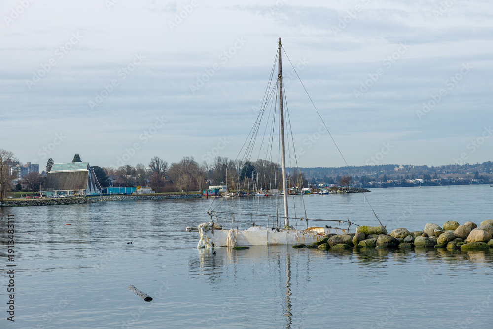 Obraz premium Sunken white sailboat with upright mast partially submerged near Vanier Park with Vancouver Maritime Museum visible on shore in background