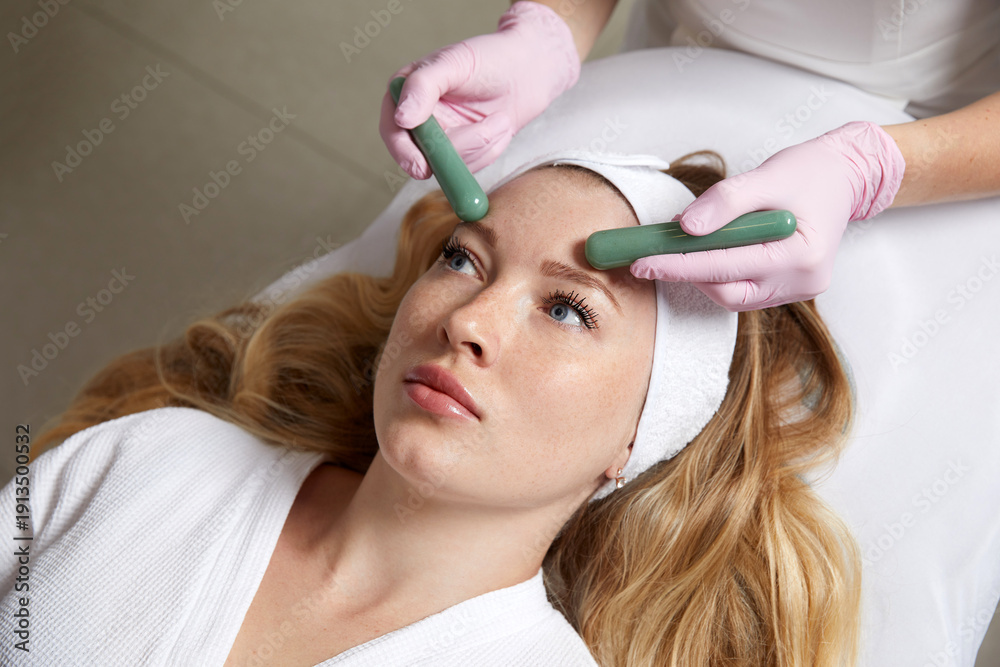 Naklejka premium Young woman receiving facial massage with stone sticks in beauty salon