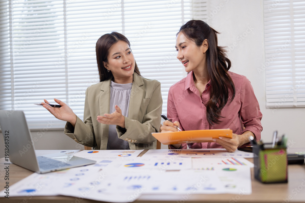 Obraz premium Two young Asian businesswomen analyzing financial reports, using calculators and discussing data on charts in a modern office.