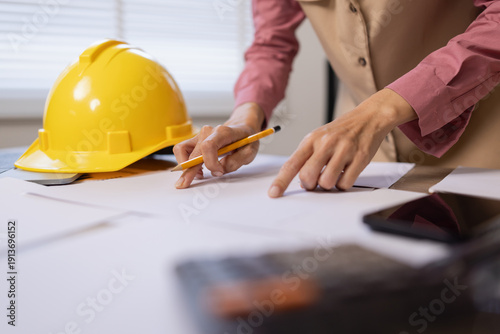 Asian female engineer or project manager in yellow hard hat working at home office, analyzing financial charts and data reports with laptop and calculator.