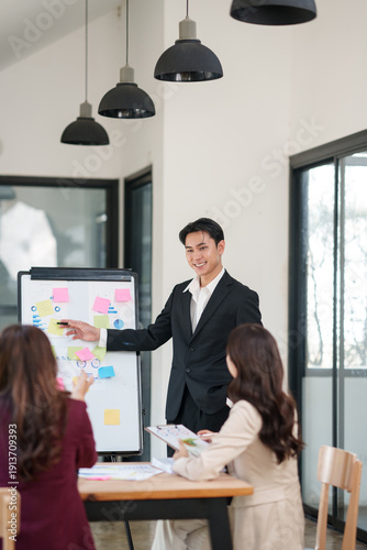 Asian businessman in a black suit giving a presentation to his female colleagues in a bright, modern office. He is pointing at a whiteboard filled with charts, graphs, and colorful sticky notes while 