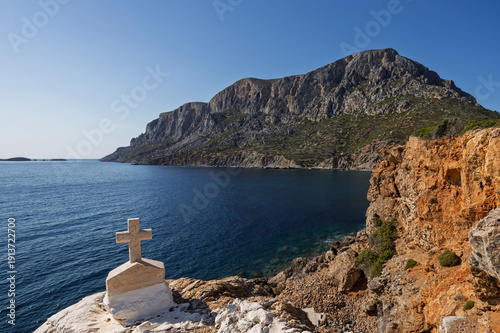 Small cross by the Aegean Sea on the rugged, rocky and mountainous island of Telendos in Greece on a sunny day.