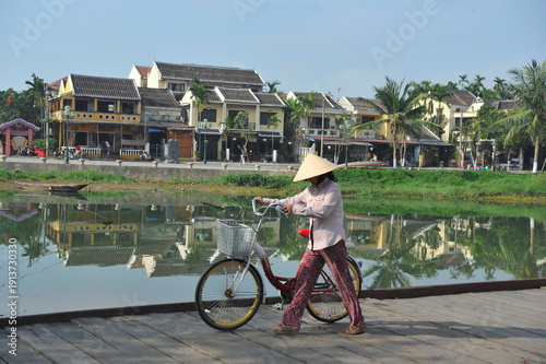 A local Vietnamese in traditional Vietnamese attire walking with a bicycle along the waterfront in the historic town of Hoi An, Vietnam. 