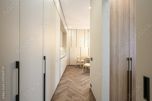 A sleek narrow hallway with light wood herringbone flooring, white cabinetry, black handles, leading to a dining area with wooden chairs and minimalist decor