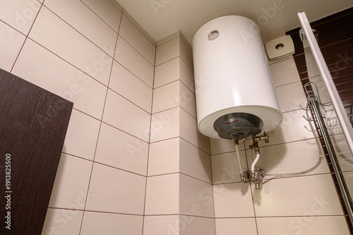 Billede på lærred White cylindrical water heater mounted on tiled wall in bathroom corner, connected with pipes, near shower cabin and dark door, clean modern setup