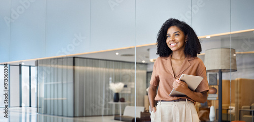 Valokuva Happy confident young African American professional woman, female entrepreneur, businesswoman looking away holding tab standing in office at work dreaming of future success