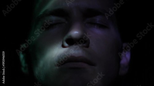 Close up of man's face, eyes closed, illuminated with red and blue light in darkness. Face lit with colorful lighting.