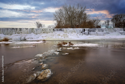 Frozen weir with icy cascades at sunset, long exposure water and winter landscape with frosted reeds and clouds.