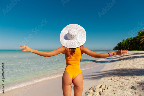 Back view of slim woman in yellow swimsuit and straw hat standing on white sandy beach towards turquoise ocean.