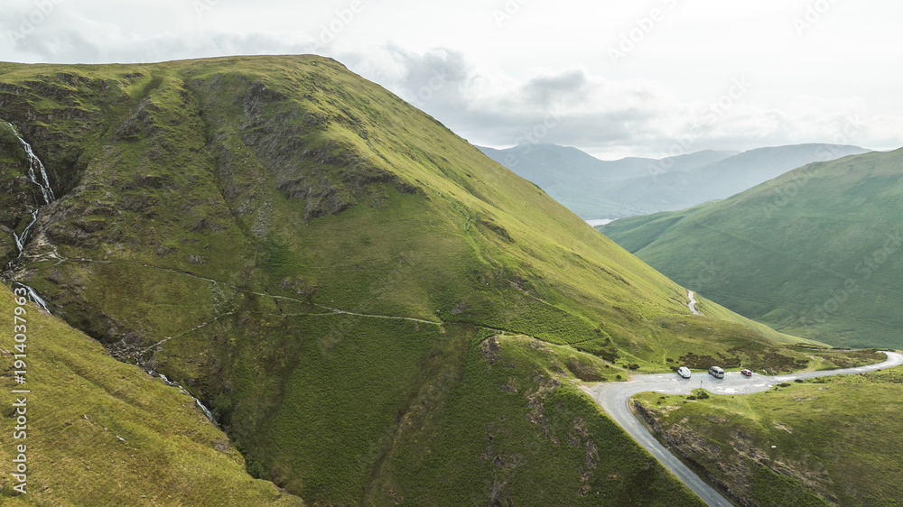 Fototapeta premium Scenic mountain pass with winding road and parked vehicles, showcasing lush green slopes and distant hazy peaks