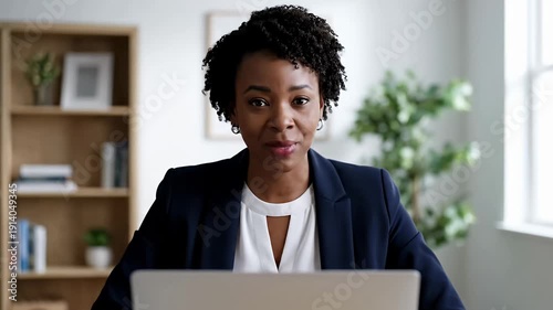 Professional woman in dark blue jacket smiles at laptop in office with bookshelf