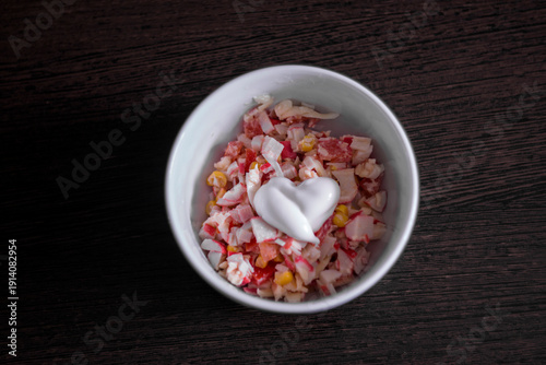 The photo shows a white bowl of crab salad with a heart-shaped mayonnaise spread in the center. The dish sits on a dark wooden table.