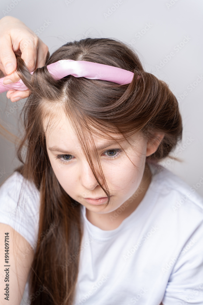 Naklejka premium Mom's hands twist curlers for daughter in white T-shirt at home on a light background in front of the mirror