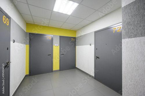 Foto A modern residential hallway with grey metal doors numbered 69–72, yellow accent wall, light grey tiled floor, white ceiling with fluorescent lighting, clean minimalist design