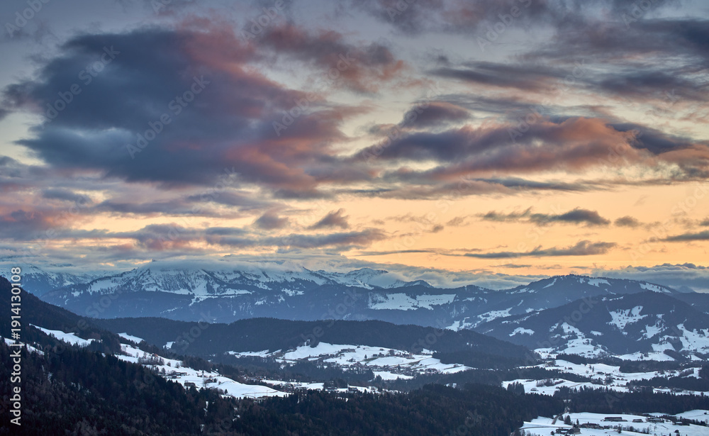 Fototapeta premium Alpine winter landscape in Bregenzer Wald at sunset with snow-covered mountains glowing in warm evening light under a dramatic sky.