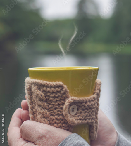 Hands holding yellow cup of hot tea in knitted cozy sleeve with misty nature background