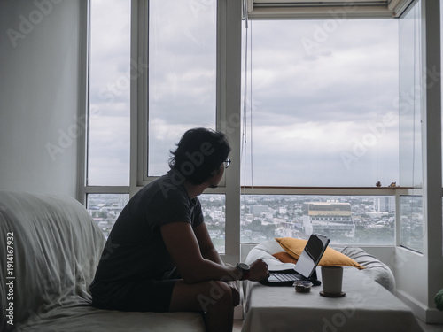 Asian man working with his digital tablet in his apartment room as work from home, feeling lonely and looking away out to the windows for city view.