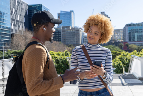 Diverse friends chatting on terrace, man with backpack holding mug, woman holding smartphone