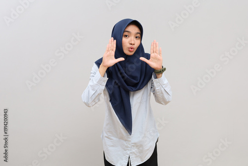 A shocked and dumbfounded young Asian woman in hijab with her palms next to her face, looking at the camera with a funny expression, reacting to something very surprising.
