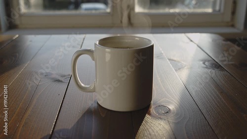 white ceramic mug filled with a hot beverage sits on a wooden table near a window. Steam rises gently from the mug, illuminated by soft sunlight streaming through the window. The scene feels cozy 