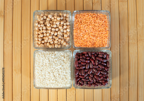 op view of assorted dry legumes and grains in glass containers on a wooden table. Chickpeas, white rice, red lentils, and red kidney beans arranged in square bowls.