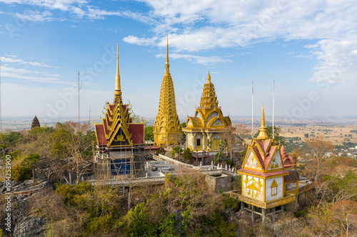 Fotografía Aerial view of ornate golden pagodas and temple structures perched atop the rocky summit of Phnom Sampov, overlooking expansive dry-season plains and rural settlements near Battambang, Cambodia