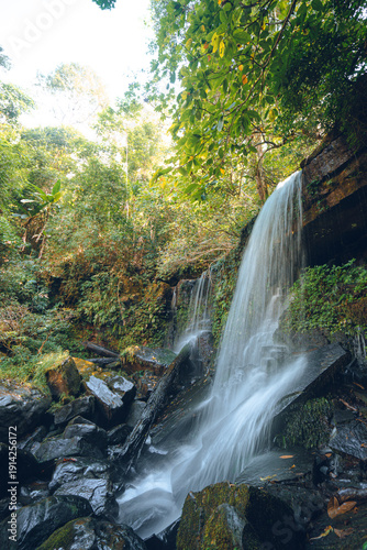 A waterfall flows down from a cliff in the forest. The water is strong in the rainy season. There are black rocks, trees, and moss all around. Idea for a natural background in soft tones.